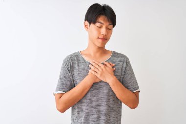 İzole edilmiş beyaz arka plan üzerinde sıradan bir tişört giyen genç Çinli adam elleri göğsünde, gözleri kapalı ve yüzünde minnet dolu bir jest ile gülümsüyor. Sağlık kavramı.