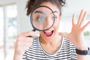 Genç Afro-Amerikalı kadın büyüteçten bakıyor çok mutlu ve heyecanlı, kazanan yüz ifadesi zaferi büyük bir gülümsemeyle kutluyor ve ellerini kaldırıyor.