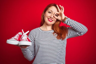 Genç, güzel, kızıl saçlı, spor ayakkabılarını kırmızı izole arka planda tutan mutlu yüzlü, gülümseyen, el pençe divan duran bir kadın.