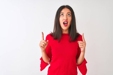İzole edilmiş beyaz arka planda duran kırmızı elbiseli genç ve güzel Çinli kadın şaşırmış ve şaşırmış bir şekilde yukarı bakıp parmaklarını göstermiş ve kollarını kaldırmıştı..