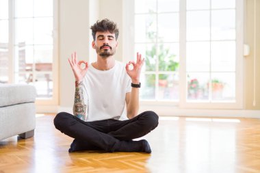 Evde rahat rahat oturup gülümseyen, gözleri kapalı meditasyon yapan genç bir adam parmaklarıyla meditasyon yapıyor. Yoga konsepti.