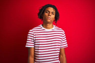 Rastalı tişörtlü genç afro adam izole edilmiş kırmızı arka planda dikiliyor. Uykulu ve yorgun görünüyor. Yorgunluktan ve akşamdan kalmalıktan yorgun..