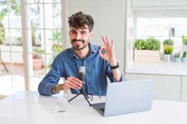 Genç adam mikrofon ve dizüstü bilgisayarla podcast kaydediyor. Parmaklarıyla imza atıyor. Mükemmel bir sembol.