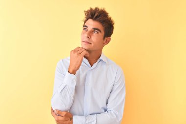 Yakışıklı genç işadamı, soyutlanmış sarı arka plan üzerinde zarif bir gömlek giyiyor ve elleri çenesinde soru sormayı düşünüyor, düşünceli bir ifade. Düşünceli bir yüzle gülümsüyor. Şüphe kavramı.