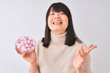 Güzel Çinli kadın izole edilmiş beyaz arka planda şekerleme kasesi tutuyor. Çok mutlu ve heyecanlı. Kazanan ifade zaferi büyük bir gülümsemeyle kutluyor ve ellerini kaldırıyor.