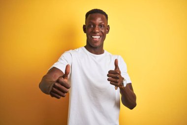 Beyaz tişörtlü genç Afro-Amerikalı adam izole edilmiş sarı arka planda duruyor. Elle olumlu jest yapıyor, gülümsüyor ve başarı için mutlu. Kazanan jest.