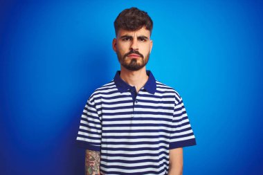 İzole edilmiş mavi arka plan üzerinde duran çizgili polo giyen genç adam şüpheli ve gergin, sorun yüzünden surat asıyor. Negatif kişi.