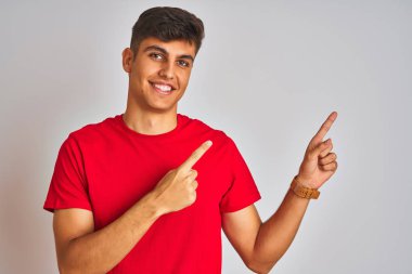 Kırmızı tişörtlü genç Hintli adam izole edilmiş beyaz arka planda duruyor gülümsüyor ve kameraya bakıyor iki eliyle ve parmaklarıyla kenarı işaret ediyor..