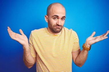Mavi izole edilmiş arka plan üzerine gündelik tişört giyen sakallı, genç ve kel bir adam. Kolları ve elleri havada, kafası karışmış bir ifade. Şüphe kavramı.