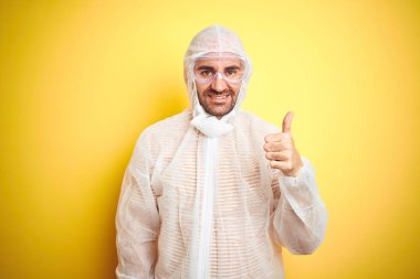 İzole edilmiş sarı arka planda boya ekipmanı ve güvenlik gözlüğü takan genç adam gülümsüyor.