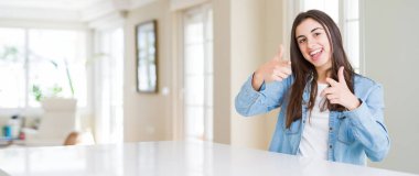 Evdeki beyaz masada oturan güzel genç bir kadının mutlu ve komik yüzüyle kamerayı işaret edişinin geniş açılı resmi. İyi enerji ve titreşimler.