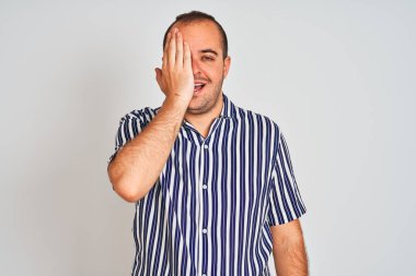Mavi çizgili gömlek giyen genç bir adam izole edilmiş beyaz arka planda duruyor bir gözünü eliyle kapatıyor, yüzünde kendine güvenen bir gülümseme ve şaşırtıcı bir duygu var..