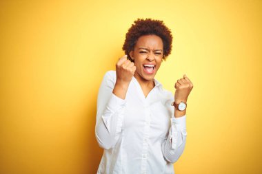 Afrika kökenli Amerikalı iş kadını, izole edilmiş sarı arka planda kollarını kaldırıp, gözlerini kapatarak başarıyı kutluyor ve şaşırıyor. Kazanan konsept.