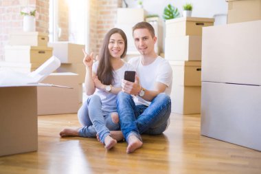 Güzel bir çift yeni evlerinde karton kutuların etrafında akıllı telefon kullanarak yerde oturuyor. Elleri ve parmakları yanda mutlu mesut işaret ediyorlar.
