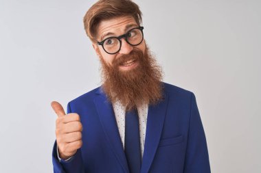Genç, kızıl saçlı, takım elbiseli ve gözlüklü bir iş adamı. İzole edilmiş beyaz arka planda gülümsüyor, gülümsüyor ve başparmağıyla yüzünü yukarı kaldırıyor..