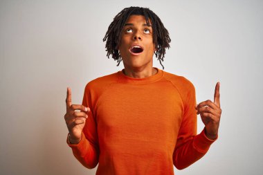 Turuncu süveter giyen Afro-Amerikalı bir adam izole edilmiş beyaz arka planda şaşkın şaşkın yukarı bakıyor ve parmaklarıyla işaret ediyor ve kollarını kaldırıyordu..