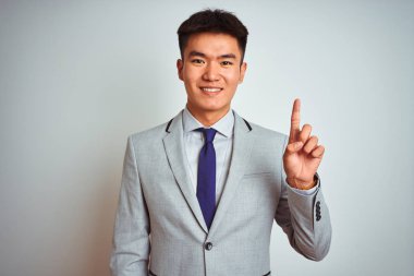 Asyalı Çinli işadamı takım elbise ve kravat takıp izole edilmiş sarı arka plan üzerinde duruyor ve bir numaralı parmağını gösterirken kendinden emin ve mutlu bir şekilde gülümsüyor..