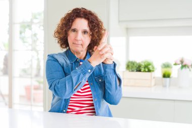 Orta yaşlı, kıvırcık saçlı, kot ceket giyen, el hareketleriyle sembolik silah tutan, ateş eden, kızgın yüzlü, orta yaşlı bir kadın.