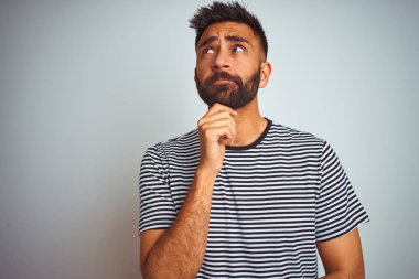 Siyah çizgili tişört giyen genç Hintli adam izole edilmiş beyaz arka planda duruyor. Elleri çenesinde soru sorarken, düşünceli bir ifade takınıyor. Düşünceli bir yüzle gülümsüyor. Şüphe kavramı.