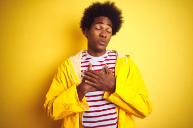 Siyah saçlı Afrikalı bir adam izole edilmiş sarı arka plan üzerinde duruyor elleri göğsünde, gözleri kapalı ve yüzünde minnet dolu bir jestle gülümsüyor. Sağlık kavramı.