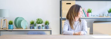 Orta yaşlı bir kadının geniş açılı fotoğrafı. Evde oturuyor, yan yana bakıyor, rahat profil pozu veriyor. Kendine güvenen bir gülümsemesi var..