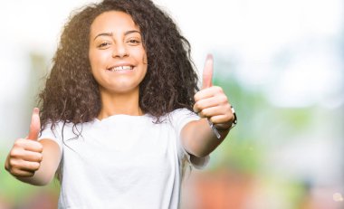 Casual beyaz t-shirt onaylama giyen kıvırcık saçlı Genç güzel kız el ile pozitif jest yapıyor gülümseyen ve başarı için mutlu yaşasın. Kamera, kazanan jest arıyorsunuz.