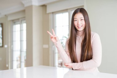 Güzel bir Asyalı kadın, beyaz masada sıradan bir kazak giyip kameraya gülümsüyor ve zafer işareti yapıyor. İki numara..