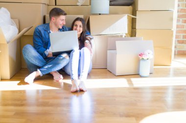 Yeni daire için mutlu gülümseyen dizüstü bilgisayar kullanarak karton kutular etrafında yeni evin katında oturan genç çift