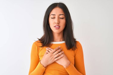 Turuncu tişört giyen genç, güzel Çinli bir kadın izole edilmiş beyaz arka plan üzerinde duruyor elleri göğsünde, gözleri kapalı ve yüzünde minnet dolu bir jest ile gülümsüyor. Sağlık kavramı.