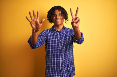 Rastgele tişörtlü Afro adam izole edilmiş sarı arka planda duruyor ve yedinci parmaklarını gösterirken kendinden emin ve mutlu bir şekilde gülümsüyor..