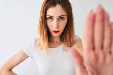 Güzel kızıl saçlı kadın, günlük tişört giyiyor, beyaz arka plan üzerinde duruyor, eli açık, dur işareti yapıyor, ciddi ve kendinden emin bir ifadeyle savunma hareketi yapıyor.