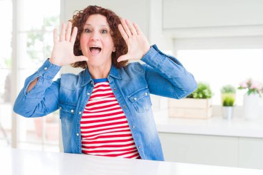 Evde kot ceket giyen kıvırcık saçlı orta yaş yaşlı kadın Gülümseyen neşeli oynarken yüzleri gösteren elleri ile bir boo gözetleme. Şaşırttı ve çıktı