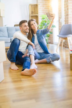 Yeni evin zemininde oturup akıllı telefon kullanarak selfie fotoğrafı çeken genç çift, yeni daireye taşındığı için mutlu bir şekilde gülümsüyor