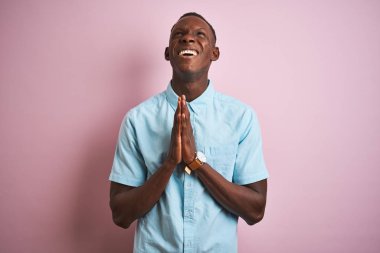 Mavi gömlekli Afro-Amerikalı bir adam pembe arka planda duruyor. Elleri bağlı yalvarıyor ve dua ediyor. Yüzünde umut ifadesi var. Af diliyor. Din kavramı.