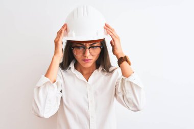 Miğfer ve gözlük takan genç, güzel bir mimar kadın izole edilmiş beyaz arka planda, acı ve migren yüzünden çaresiz ve stresli bir baş ağrısı çekiyor. Ellerini başının üstüne koy..