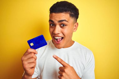 Genç Brezilyalı müşteri kredi kartı tutuyor. İzole edilmiş sarı arka planda duruyor. Elle ve parmakla göstererek çok mutlu.