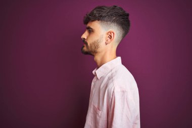 Dövmeli, pembe tişörtlü genç bir adam. Mor arka planda duruyor. Yan tarafa bakıyor..