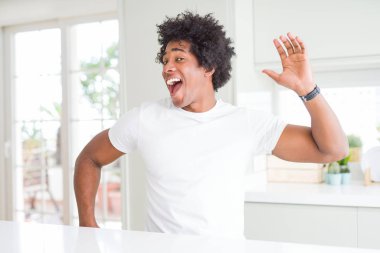 Genç Afro-Amerikalı adam, günlük beyaz tişörtler giyip evde oturmuş mutlu ve neşeli dans ediyor, gülümsüyor, rahat ve kendinden emin bir şekilde müzik dinliyor.