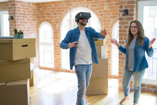 Young couple moving to a new house having fun playing with virtual reality glasses around cardboard boxes