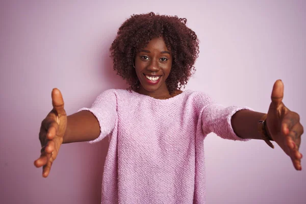 Young african afro woman wearing sweater standing over isolated pink background looking at the camera smiling with open arms for hug. Cheerful expression embracing happiness.