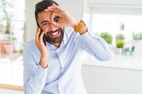 Handsome hispanic business man having a conversation talking on smartphone with happy face smiling doing ok sign with hand on eye looking through fingers