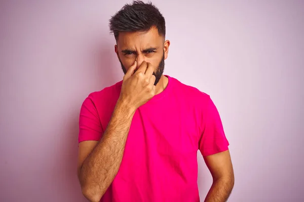 年轻的印度人穿着T恤站在孤立的粉色背景上 闻到一股难闻而恶心的气味 用手指捂住鼻子屏住呼吸 臭味概念 — 图库照片