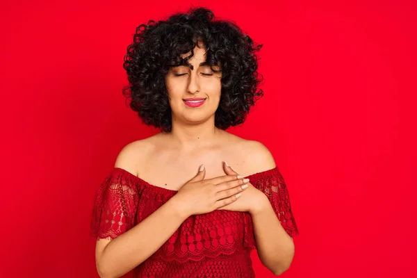 閉じた目と顔に感謝のジェスチャーで胸に手で笑みを浮かべて孤立した赤い背景の上にカジュアルなドレスを着て巻き毛を持つ若いアラブの女性 健康コンセプト — ストック写真
