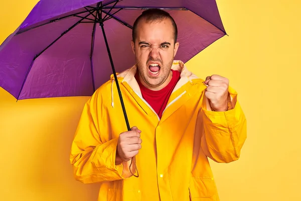 Young man wearing raincoat holding purple umbrella standing over isolated yellow background annoyed and frustrated shouting with anger, crazy and yelling with raised hand, anger concept