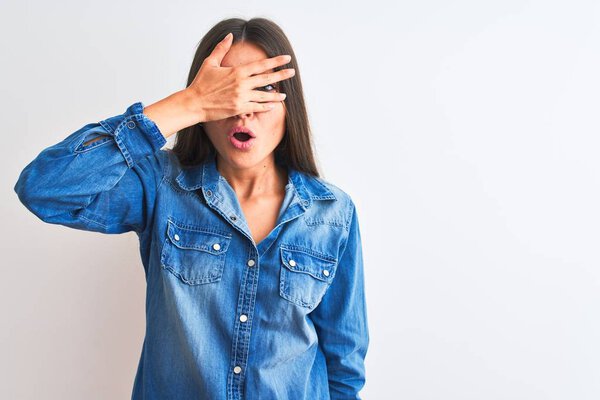 Молодая красивая женщина в джинсовой рубашке, стоящая на изолированном белом фоне, заглядывая в шокирующее лицо и глаза рукой, глядя сквозь пальцы со смущенным выражением лица
.
