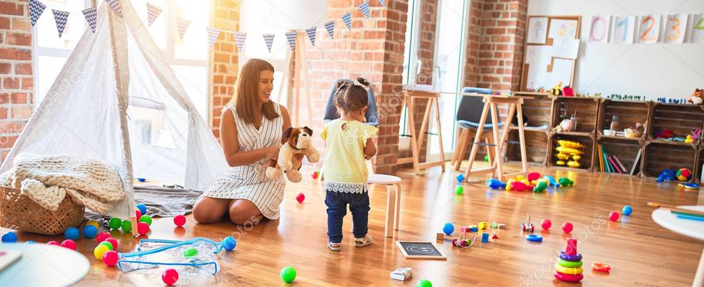 Young beautiful teacher playing with dog doll and toddler holding jar of chocolate balls at kindergarten