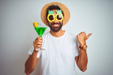 Yazın komik giyinen Hintli adam izole edilmiş beyaz arka planda kokteyl içiyor ve gülümseyen bir gülümsemeyle baş parmağıyla onu işaret ediyor.