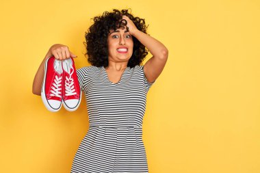 Kıvırcık saçlı genç bir Arap kadın izole edilmiş sarı arka planda spor ayakkabılarını tutuyor. Elleri başının üstünde, utanç ve şaşkınlık dolu, sinirli ve hayal kırıklığına uğramış bir halde. Hata için korku ve üzüntü.