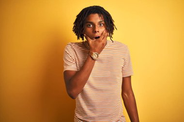 Sarı arka plan üzerinde duran çizgili t-shirt giyen Afro bir adam inançsızlıktan, şaşkınlıktan ve elleri çenesinde olan şaşkınlıktan büyülenmiş görünüyor.