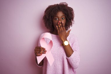 Genç Afrikalı Afro kadın, elinde kanserli kurdele, pembe arka planda duruyor. Ağzını kapatıyor. Yanlışlıkla utancından ağzı sarsılıyor.